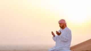 Man in traditional attire kneeling in desert, praying at sunset with warm glow on sand.