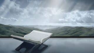 Open Quran with Arabic text on a stand, set against green hills and cloudy sky.