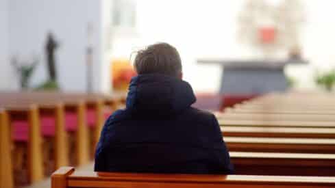 Person in dark jacket sits alone on pew in bright, empty church, altar visible in distance.