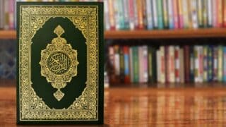 Decorative Quran with ornate gold design on dark cover, wooden surface, blurred colorful bookshelf.