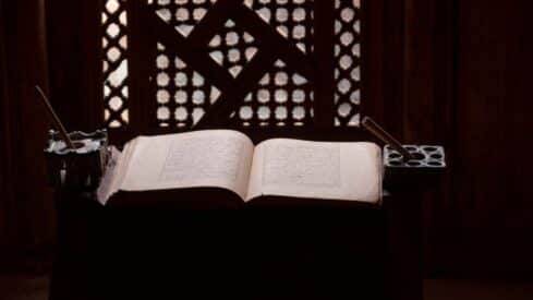 Open Quran on a stand with inkwells and pens, against a patterned wooden lattice window.