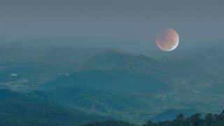 Landscape with forested hills, large moon, and twilight sky, misty serene atmosphere.