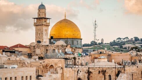 Cityscape with Dome of the Rock's gold dome, historical buildings, antennas, and cloudy sky.