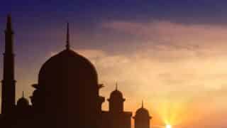 Silhouette of a mosque with domes and spires against a transitioning orange-blue sky at sunset.