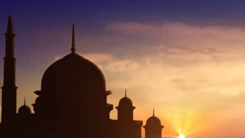 Silhouette of a mosque with domes and spires against a transitioning orange-blue sky at sunset.