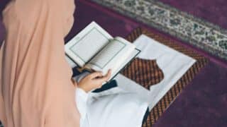 Person in headscarf reading Quran book on prayer rug with dark decorative carpet.