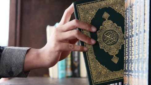 Hand reaching for a green Quran book with gold designs and Arabic script, shelves in background.