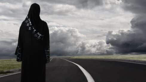 Person in black garment with patterns on road under cloudy sky, fields on sides.