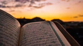 Open Quran with Arabic text, sunset behind, warm sky hues, silhouetted structures below.