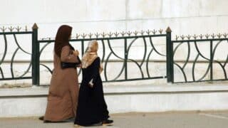 Two individuals, in long coats and headscarves, walk near an ornate metal fence on a sidewalk.