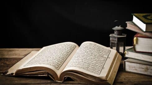 Open Quran with Arabic script, wooden surface, stack of closed books, small lantern, dark background.