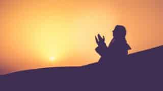 Silhouette of a person praying against an orange sky during sunrise or sunset.