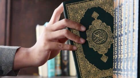 Hand removing ornate Quran with gold Arabic script from shelf.