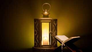 Decorative lantern lit by a candle, next to an open book with prayer beads on a stand.