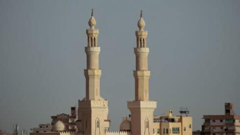 Two ornate beige minarets of a mosque against a clear sky with buildings in the background.