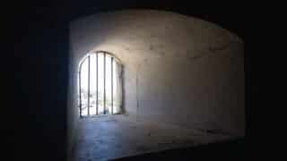 Small room with curved ceiling and barred window, rough stone surface, light entering.