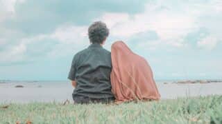 Two people seated on grass facing the sea, under a partly cloudy sky, with rocks visible.