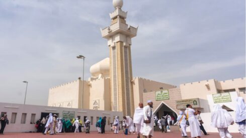 People in white garments gather near a light-colored jirana mosque with minaret under clear blue sky.