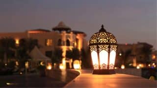 Ornate lantern glowing warmly on a smooth surface; building and palm trees in background.