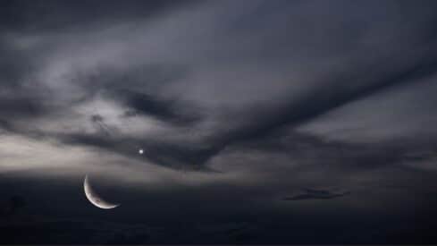 Crescent moon and bright star or planet in a dark, cloudy nighttime sky.