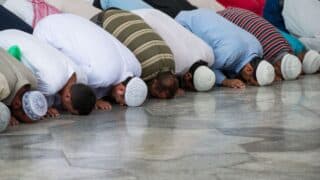 Men kneeling in prayer on polished stone, wearing caps and colorful garments.