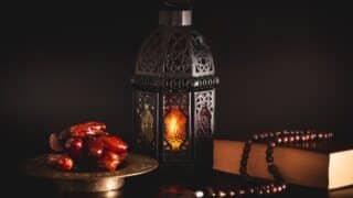 A dark lantern glows warmly beside dates, a wooden bead string, and a book on a dark background.