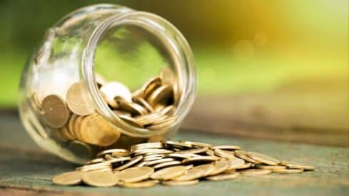 Glass jar tipped, gold coins spilling on wood, blurred green background.