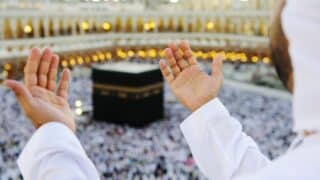 Person with hands raised in prayer facing the Kaaba, surrounded by a crowd and lit mosque.