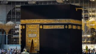 Kaaba surrounded by people in a mosque, with scaffolding in the background and gold embroidery.