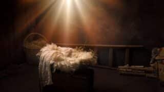 Dimly lit rustic room with a straw-lined manger, basket, and wooden ladder. Warm light above.