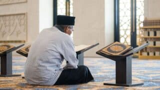 Person reading Quran on wooden stand in ornate room with carpet, bookshelves, and decorated walls.