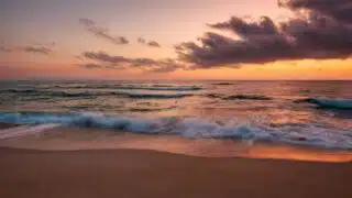 Beach at sunset with waves on sandy shore, clouds reflecting warm hues in the sky.