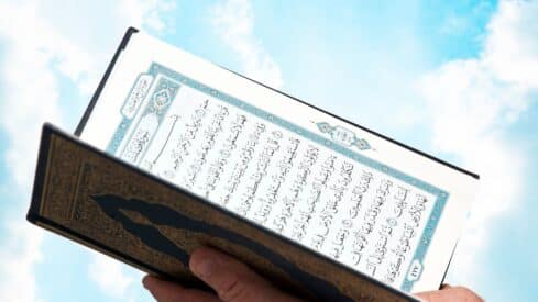 Quran book with Arabic text held against a blue sky with scattered clouds