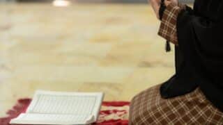 Person in plaid kneeling with prayer book and beads on patterned rug.