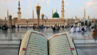 Open Quran book, mosque with green dome and minarets, people in courtyard.