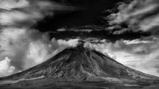 Black and white volcanic scene with plume, dramatic clouds in the sky, rugged surrounding terrain.