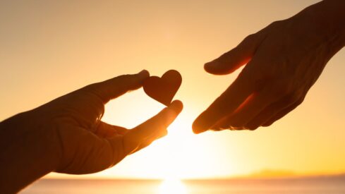 Hands forming a heart with fingers against an orange sunset, symbolizing love and happiness in nature