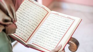 كتاب مفتوح يُظهر صفحات مكتوبة باللغة العربية مع نصوص دينية، يُمسك به شخص يرتدي حجاب. الخلفية غير واضحة، مما يركز الانتباه على الكتاب.