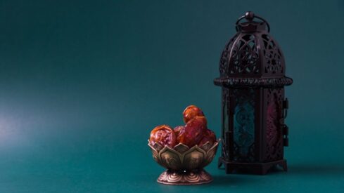 A decorative bowl filled with dates placed next to an ornate lantern against a teal background, showcasing a blend of traditional Middle Eastern elements.