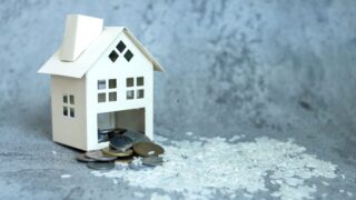 Model house made of paper with a slot for coins, surrounded by scattered coins and white rice on a textured gray background. Represents savings, investment, and financial planning in real estate.